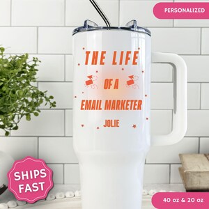 May include: White insulated tumbler with a handle, featuring the text "THE LIFE OF A EMAIL MARKETER" in orange. The name "JOLIE" is below the text. The tumbler has a black straw and a clear lid. The tumbler is available in 40 oz and 20 oz sizes.
