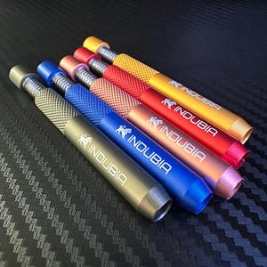 May include: Five cylindrical metal tools with knurled grips and threaded ends, arranged diagonally. Each tool features the brand name INDUBIA and a lion logo printed in white. The colors are olive green, blue, copper, red, and gold, all metallic.