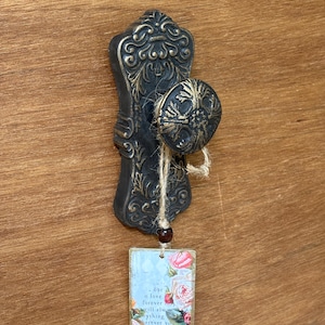 May include: A dark bronze-toned metal door knob with an ornate, embossed backplate. A piece of twine is tied around the knob, suspending a small rectangular tag featuring a floral print and the text: our love forever will always be everything wherever you are, bless and you.