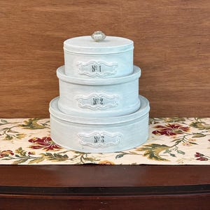 May include: A stack of three round, light blue decorative boxes with lids. Each box is labeled with a number: "No. 1", "No. 2", and "No. 3". The top box has a crystal knob. The boxes are on a floral runner.