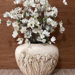 May include: A cream-colored ceramic vase with a rounded shape and a textured relief pattern of stylized flowers and stems. The vase is filled with a large arrangement of white dogwood blossoms featuring thin brown branches and small green leaves.