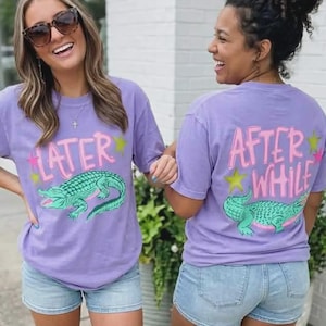 May include: Two women wearing matching lavender t-shirts and denim shorts. The shirts feature a green alligator graphic and the text "LATER" on the front and "AFTER WHILE" on the back, with pink and yellow accents.