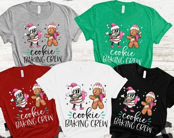 May include: A collection of t-shirts in various colors, including gray, green, red, white, and black. Each shirt features a cartoon design of a gingerbread man and a cookie character wearing Santa hats, holding drinks, and the text "cookie BAKING CREW".
