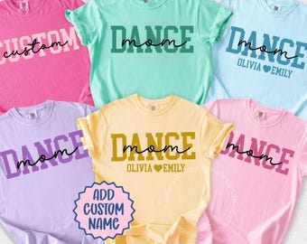 以下が含まれることがあります： ピンク、緑、青など、さまざまな色の半袖Tシャツが6枚。各シャツには、太字で「DANCE mom」または「CUSTOM」の文字がプリントされています。名前などの追加テキストが記載されているシャツもあります。カジュアルなスタイル。