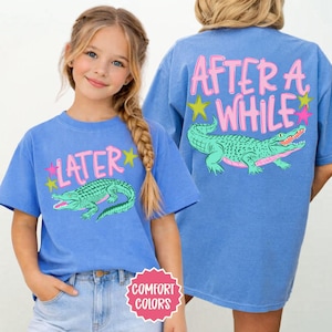 May include: Blue t-shirts with a cartoon alligator design. One shirt has "LATER" and the other has "AFTER A WHILE" in pink text. Both shirts have a green alligator with pink accents and star graphics.