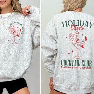 K&ouml;nnte beinhalten: Hellgraues Sweatshirt mit wei&szlig;em Saum. Auf der Vorderseite befindet sich eine rot-wei&szlig;e Grafik von Santa in einem Cocktailglas mit dem Text "FULL OF HOLIDAY SPIRIT". Auf der R&uuml;ckseite steht "HOLIDAY CHEER" und "COCKTAIL CLUB".