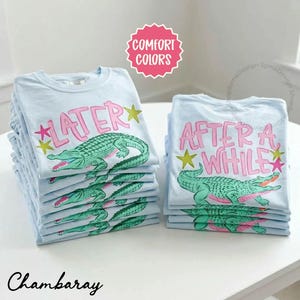 May include: Stacks of light blue t-shirts with a cartoon alligator design. One stack has the word "LATER" in pink with a green alligator, and the other has "AFTER A WHILE". The shirts are on a white surface.