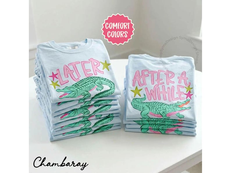 May include: Stacks of light blue t-shirts with a cartoon alligator design. One stack has the word "LATER" in pink with a green alligator, and the other has "AFTER A WHILE". The shirts are on a white surface.
