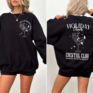 K&ouml;nnte beinhalten: Schwarzes Sweatshirt mit wei&szlig;er Grafik von Santa Claus in einem Cocktailglas. Auf der Vorderseite steht "FULL OF HOLIDAY SPIRIT". Auf der R&uuml;ckseite steht "HOLIDAY CHEER COCKTAIL CLUB MAKING SPIRITS BRIGHT".