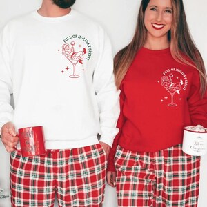 K&ouml;nnte beinhalten: Wei&szlig;e und rote Sweatshirts mit einem festlichen Motiv. Das Design zeigt den Weihnachtsmann in einem Martini-Glas mit dem Text "Full of Holiday Spirit". Die Models tragen rot-gr&uuml;n karierte Pyjamahosen.