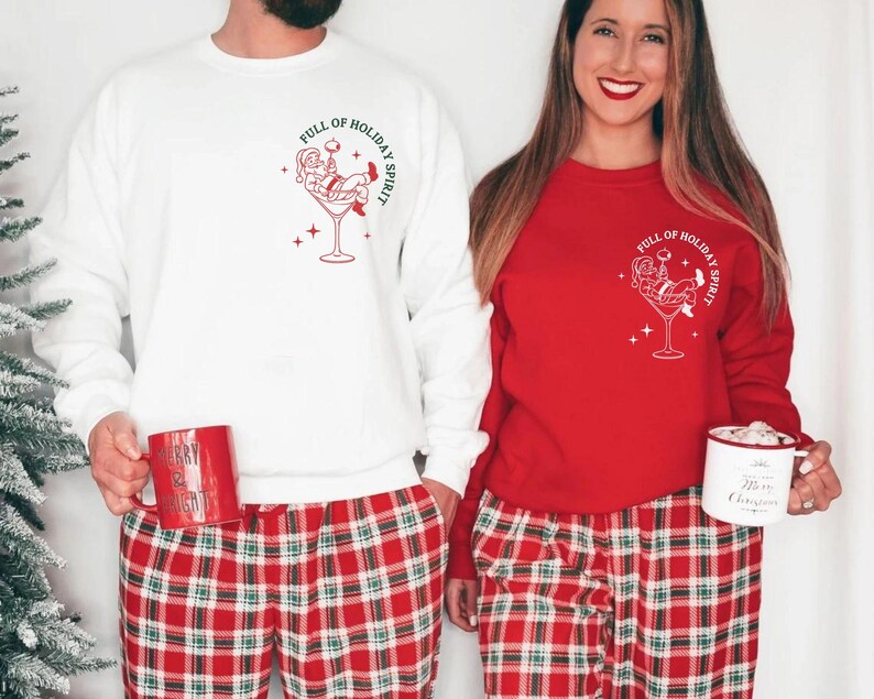 K&ouml;nnte beinhalten: Wei&szlig;e und rote Sweatshirts mit einem festlichen Motiv. Das Design zeigt den Weihnachtsmann in einem Martini-Glas mit dem Text "Full of Holiday Spirit". Die Models tragen rot-gr&uuml;n karierte Pyjamahosen.