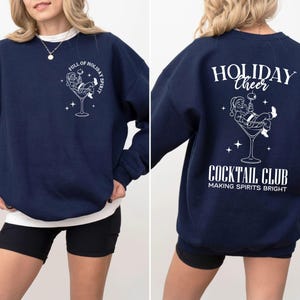 K&ouml;nnte beinhalten: Marineblaues Sweatshirt mit wei&szlig;er Grafik von Santa Claus in einem Cocktailglas. Auf der Vorderseite steht "FULL OF HOLIDAY SPIRIT". Auf der R&uuml;ckseite steht "HOLIDAY CHEER COCKTAIL CLUB MAKING SPIRITS BRIGHT."