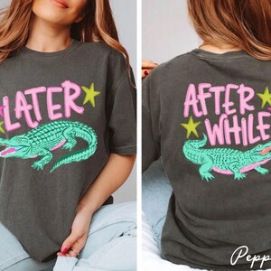 May include: Dark gray t-shirt with a front graphic that says "LATER" above a teal alligator illustration. The back of the shirt says "AFTER WHILE" above the same alligator design. Pink and yellow stars accent the text.