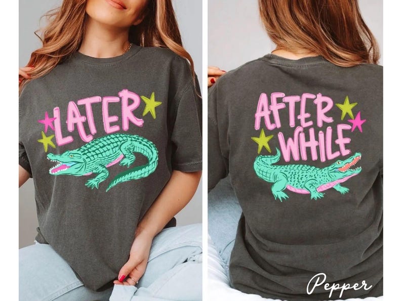 May include: Dark gray t-shirt with a front graphic that says "LATER" above a teal alligator illustration. The back of the shirt says "AFTER WHILE" above the same alligator design. Pink and yellow stars accent the text.