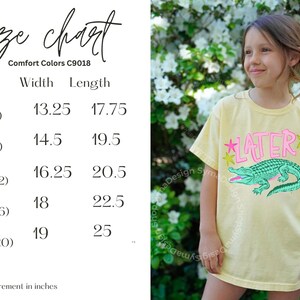 May include: A light yellow t-shirt with the word "LATER" in pink and a green alligator graphic. The shirt has a size chart with measurements in inches. The model is wearing a size small.