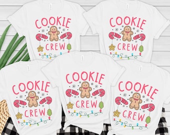 May include: White t-shirts with the text "COOKIE CREW" in pink, featuring a gingerbread man, candy canes, stars, Christmas trees, and string lights. A festive design for the holiday season.