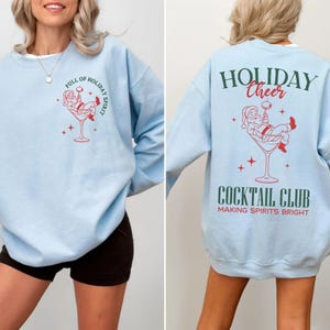 K&ouml;nnte beinhalten: Hellblaues Sweatshirt mit roter und wei&szlig;er Grafik von Santa Claus in einem Cocktailglas. Auf der Vorderseite steht "FULL OF HOLIDAY SPIRIT". Auf der R&uuml;ckseite steht "HOLIDAY CHEER COCKTAIL CLUB MAKING SPIRITS BRIGHT".
