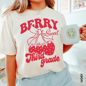 May include: Ivory-colored t-shirt with a red graphic that reads "Berry Sweet Third Grade." The graphic features a bow and strawberries. A white mug with a smiley face is held in the hand.