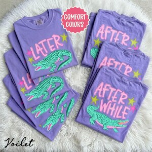 May include: Several purple t-shirts with the words "LATER" and "AFTER WHILE" in pink, along with a green alligator graphic. The shirts have a crew neck and short sleeves. The text "Comfort Colors" is in a pink circle.