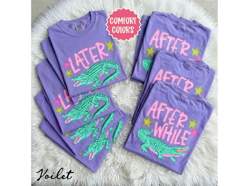 May include: Several purple t-shirts with the words "LATER" and "AFTER WHILE" in pink, along with a green alligator graphic. The shirts have a crew neck and short sleeves. The text "Comfort Colors" is in a pink circle.