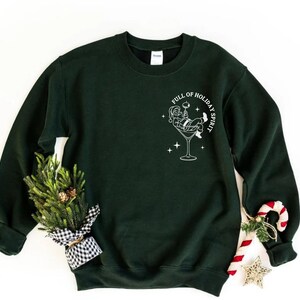 K&ouml;nnte beinhalten: Ein dunkelgr&uuml;nes Sweatshirt mit einer wei&szlig;en Grafik von Santa Claus in einem Martini-Glas, mit dem Text "FULL OF HOLIDAY SPIRIT". Ein kleiner Weihnachtsbaum und eine Zuckerstange sind an der Seite.