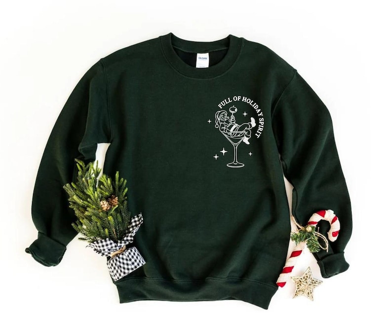 K&ouml;nnte beinhalten: Ein dunkelgr&uuml;nes Sweatshirt mit einer wei&szlig;en Grafik von Santa Claus in einem Martini-Glas, mit dem Text "FULL OF HOLIDAY SPIRIT". Ein kleiner Weihnachtsbaum und eine Zuckerstange sind an der Seite.