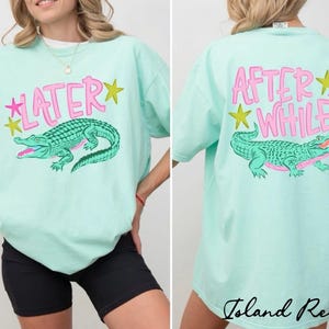 May include: Mint green t-shirt with a pink and green alligator graphic. The front of the shirt says "LATER" with a pink star and yellow star. The back says "AFTER WHILE" with a pink star and yellow star.