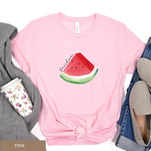 May include: A pale pink t-shirt with a watercolour illustration of a watermelon slice. The watermelon has red flesh, black seeds, and a green rind. The word "freedom" is written in cursive above the watermelon.