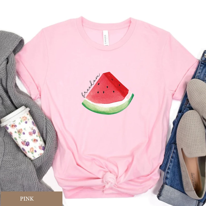 May include: A pale pink t-shirt with a watercolour illustration of a watermelon slice. The watermelon has red flesh, black seeds, and a green rind. The word "freedom" is written in cursive above the watermelon.