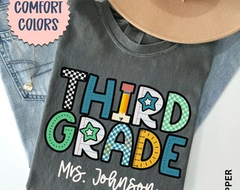 May include: A gray t-shirt with the words "THIRD GRADE" in colorful block letters, a pencil, and a ruler graphic. The text "Mrs. Johnson" is below the grade level. A tan hat and blue jeans are in the background.