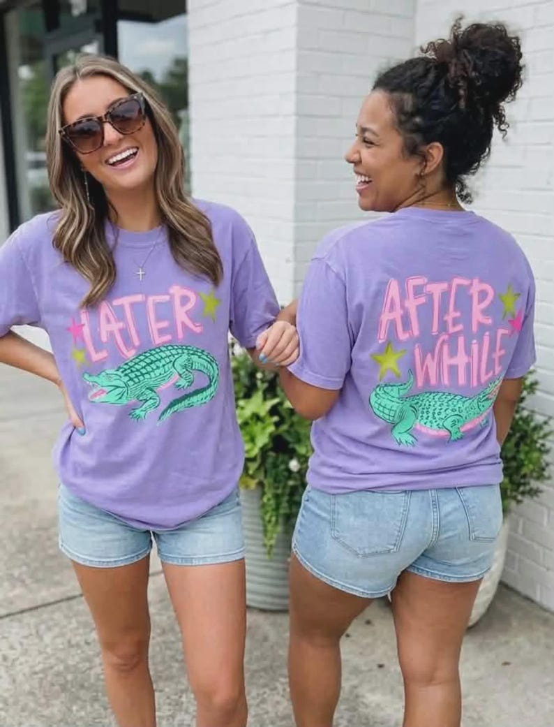 May include: Two women wearing matching lavender t-shirts and denim shorts. The shirts feature a green alligator graphic and the text "LATER" on the front and "AFTER WHILE" on the back, with pink and yellow accents.