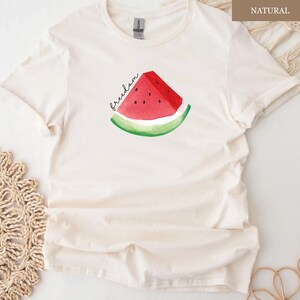 May include: Cream-coloured t-shirt featuring a watercolour watermelon slice with the word "freedom" in black script. The watermelon has red flesh, black seeds, and a green rind. The shirt is made of a soft, light-coloured fabric.