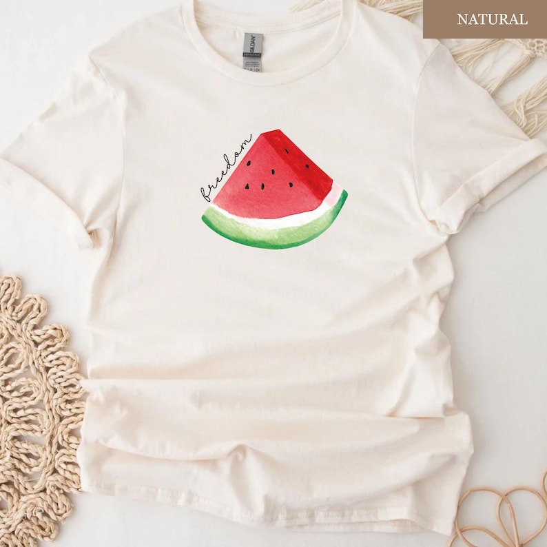 May include: Cream-coloured t-shirt featuring a watercolour watermelon slice with the word "freedom" in black script. The watermelon has red flesh, black seeds, and a green rind. The shirt is made of a soft, light-coloured fabric.
