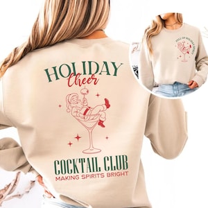 K&ouml;nnte beinhalten: Beiges Sweatshirt mit festlichem Design. Auf der R&uuml;ckseite befindet sich eine Illustration des Weihnachtsmanns in einem Martini-Glas mit den Worten "HOLIDAY CHEER" und "COCKTAIL CLUB". Auf der Vorderseite befindet sich eine kleinere Version des Designs und die Worte "FULL OF HOLIDAY SPIRIT".