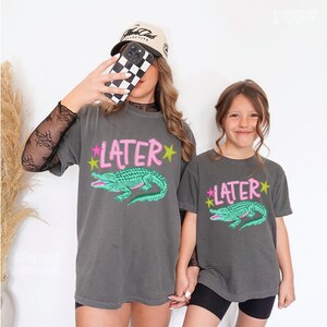 May include: Two gray t-shirts with the word "LATER" in pink and a green alligator graphic. The shirts are worn by two people. One person is wearing a black and white checkered phone case and a baseball cap.