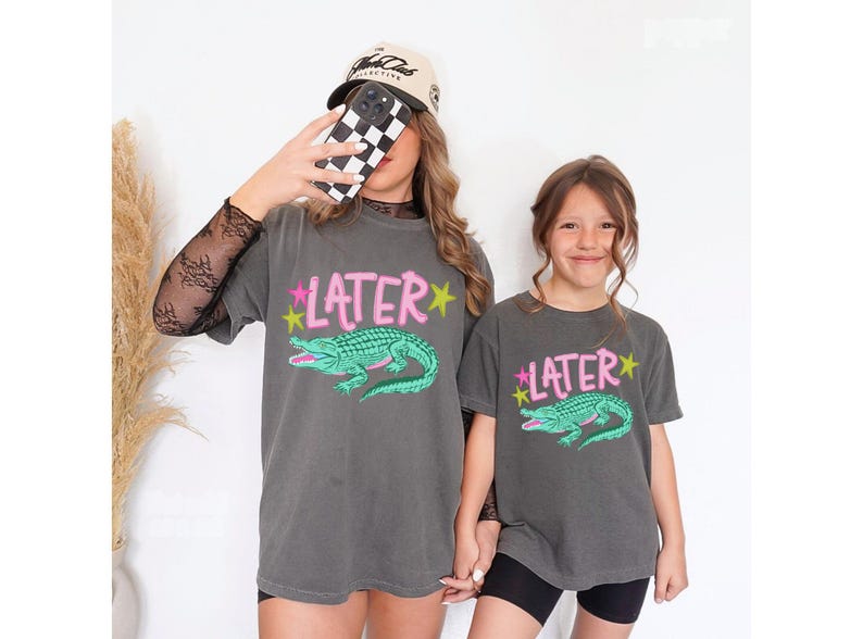 May include: Two gray t-shirts with the word "LATER" in pink and a green alligator graphic. The shirts are worn by two people. One person is wearing a black and white checkered phone case and a baseball cap.