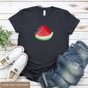 May include: Dark grey heather t-shirt featuring a watercolour watermelon slice and the word "freedom". The image includes white trainers and blue jeans, suggesting a relaxed, summery outfit.