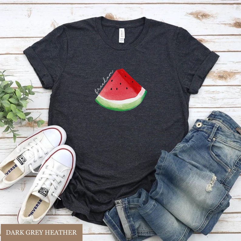 May include: Dark grey heather t-shirt featuring a watercolour watermelon slice and the word "freedom". The image includes white trainers and blue jeans, suggesting a relaxed, summery outfit.
