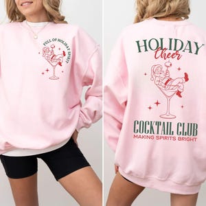 K&ouml;nnte beinhalten: Hellrosa Sweatshirt mit festlichem Design. Auf der Vorderseite befindet sich ein Weihnachtsmann in einem Martini-Glas mit dem Text "FULL OF HOLIDAY SPIRIT". Auf der R&uuml;ckseite stehen "HOLIDAY CHEER" und "COCKTAIL CLUB" mit einem Weihnachtsmann-Motiv.