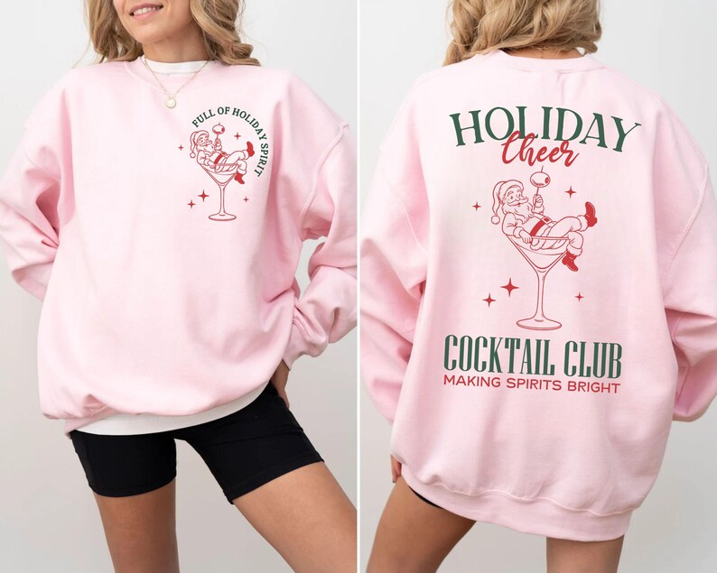 K&ouml;nnte beinhalten: Hellrosa Sweatshirt mit festlichem Design. Auf der Vorderseite befindet sich ein Weihnachtsmann in einem Martini-Glas mit dem Text "FULL OF HOLIDAY SPIRIT". Auf der R&uuml;ckseite stehen "HOLIDAY CHEER" und "COCKTAIL CLUB" mit einem Weihnachtsmann-Motiv.