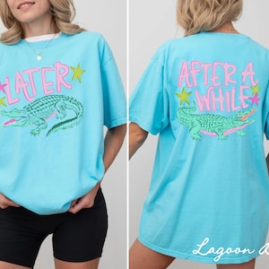May include: Lagoon blue t-shirt with a front graphic of a green alligator and the word "LATER" in pink. The back of the shirt features the words "AFTER A WHILE" and a matching alligator graphic. The shirt has a relaxed fit.