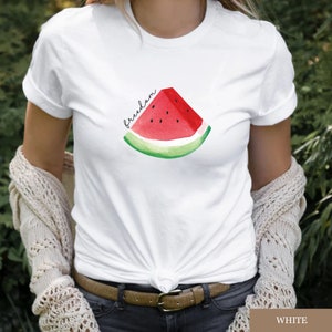 May include: White t-shirt with a watercolour watermelon slice design and the word "freedom". The watermelon has red flesh, black seeds, and a green rind. A casual, everyday wear item.