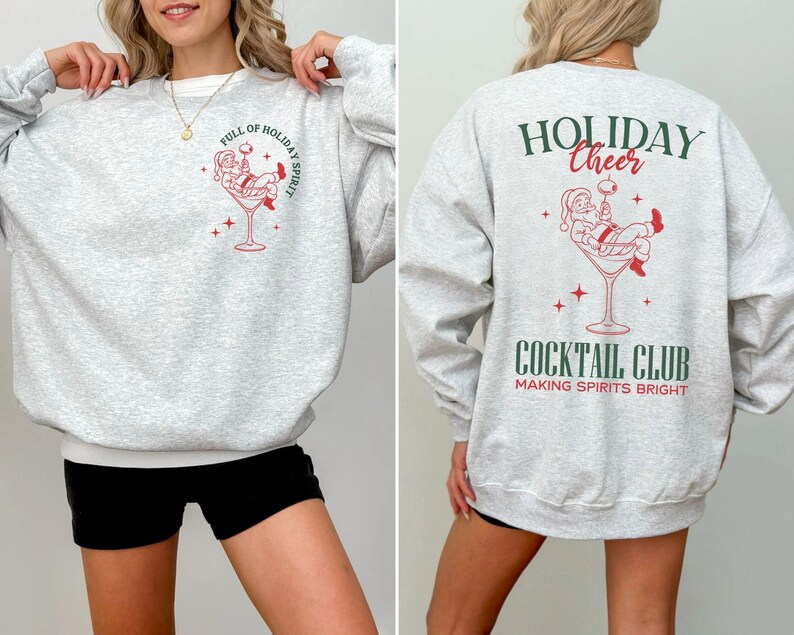 K&ouml;nnte beinhalten: Hellgraues Sweatshirt mit wei&szlig;em Saum. Auf der Vorderseite befindet sich eine rot-wei&szlig;e Grafik von Santa in einem Cocktailglas mit dem Text "FULL OF HOLIDAY SPIRIT". Auf der R&uuml;ckseite steht "HOLIDAY CHEER" und "COCKTAIL CLUB".