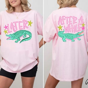 May include: Pink t-shirt with a front graphic of a teal alligator and the word "LATER" in pink. The back of the shirt features the phrase "AFTER A WHILE" and a matching alligator graphic. The shirt has a relaxed fit.