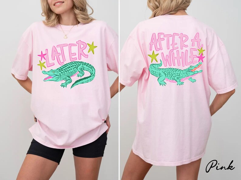May include: Pink t-shirt with a front graphic of a teal alligator and the word "LATER" in pink. The back of the shirt features the phrase "AFTER A WHILE" and a matching alligator graphic. The shirt has a relaxed fit.