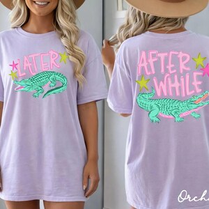 May include: A light purple t-shirt with a front and back design. The front features the word "LATER" above a green alligator graphic. The back says "AFTER WHILE" above a matching alligator. Both designs include star accents.