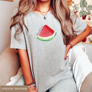May include: A grey t-shirt with a watercolour watermelon slice design and the word "freedom". The watermelon is red, green, and black. This casual t-shirt is suitable for everyday wear.