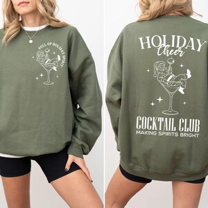 K&ouml;nnte beinhalten: Olivgr&uuml;nes Sweatshirt mit wei&szlig;er Grafik von Santa Claus in einem Martini-Glas. Auf der Vorderseite steht "FULL OF HOLIDAY SPIRIT". Auf der R&uuml;ckseite steht "HOLIDAY CHEER COCKTAIL CLUB MAKING SPIRITS BRIGHT".