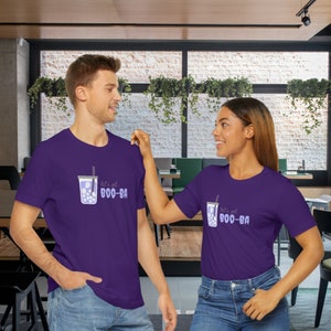 May include: Two people wearing matching purple t-shirts. Each shirt features a graphic of a bubble tea with the text "let's get BOO-BA" in light purple. The bubble tea graphic includes a straw and boba pearls.