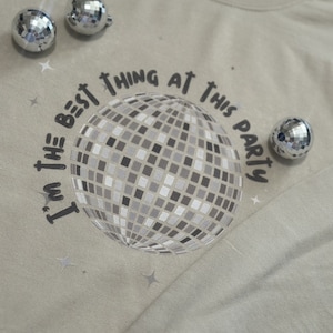 May include: A light tan long-sleeve shirt with a disco ball graphic and the text "I'm the best thing at this party." Three small disco ball ornaments are scattered around the shirt.